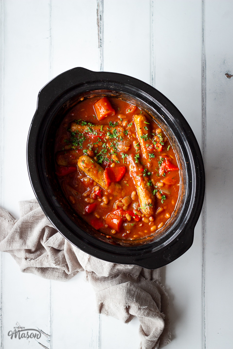 An angled top down view of a slow cooked sausage casserole with beans in a slow cooker on a white wood effect backdrop. There is also a light brown linen napkin scrunched up in the background.