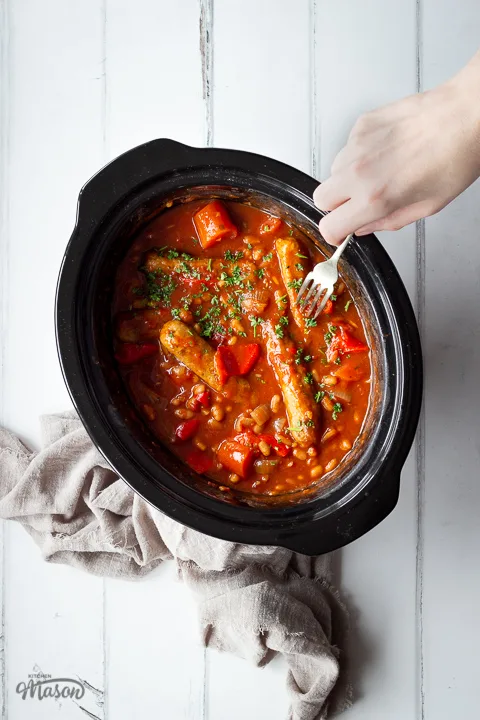 An angled top down view of a slow cooked sausage casserole with beans in a slow cooker on a white wood effect backdrop. There is also a light brown linen napkin scrunched up in the background and a person reaching into the shot with a fork.
