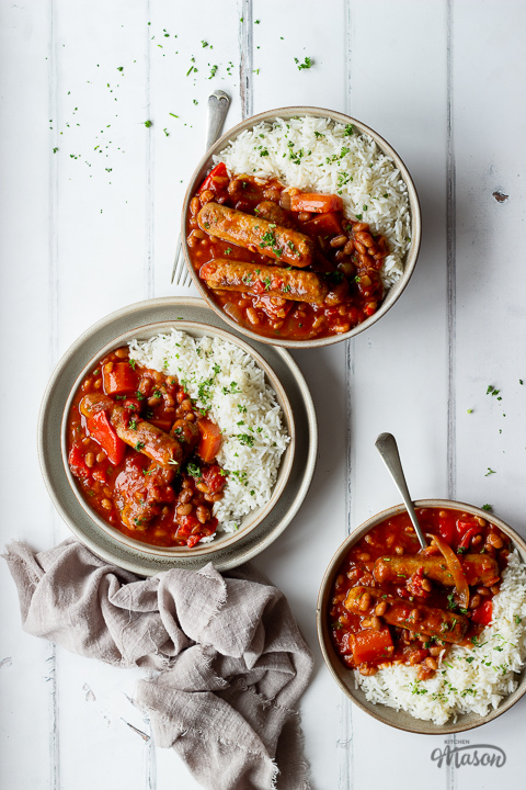 A top down view of 3 bowls filled with slow cooker sausage casserole with beans and rice. Set over a white wood effect backdrop, there are also some forks, scattered chopped parsley and a light brown linen napkin in the background.