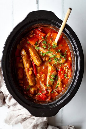 A top down view of a slow cooker filled with sausage casserole with beans. Set against a white wood effect backdrop there is a wooden spoon in the bowl and a light brown linen napkin to the side.