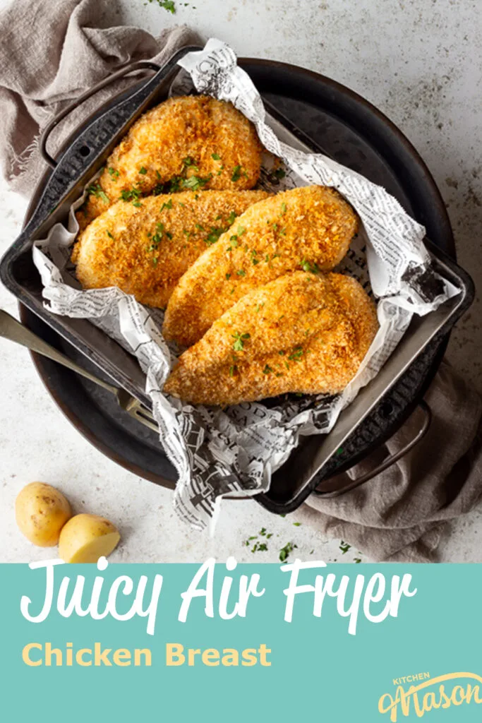 4 x Juicy air fried chicken breasts with a crispy golden crumb coating in a baking tray lined with baking paper. Set over a grey pewter tray on a light brown linen napkin. There is also a fork on the side and some scattered potatoes and chopped parsley in the background. A text overlay says "Juicy Air Fryer Chicken Breast"