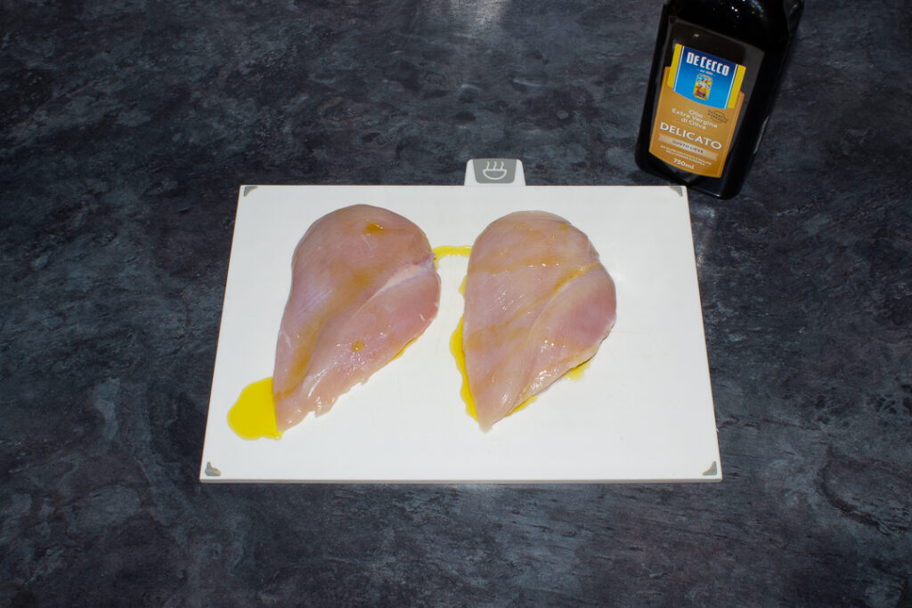 2 x large chicken breasts on a white chopping board coated in olive oil.