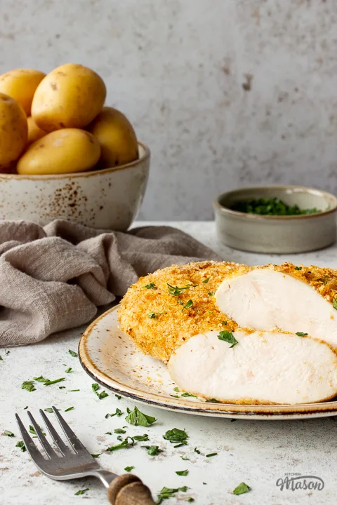 A front view of a sliced air fried chicken breast, showing the juicy inside. Set on a mottled white plate over a white mottled background. There is also a bowl of potatoes, a wooden handled fork, a pot of parsley and a light brown linen napkin in the background.