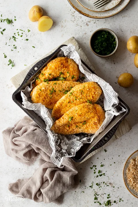 4 juicy air fried chicken breasts coated in golden breadcrumbs set in a baking tin lined with paper over a white chopping board. There are also potatoes, plates, scattered parsley, a light brown linen napkin and a small pot of parsley in the background.