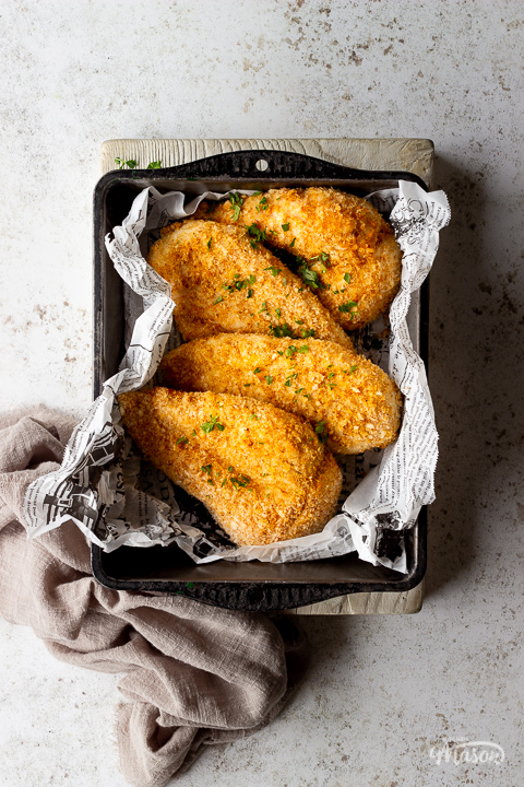 4 x juicy air fried chicken breasts with a crispy golden coating in a baking tray lined with scrunched up paper. Set over a white mottled backdrop on a white board with a light brown linen napkin to the side.