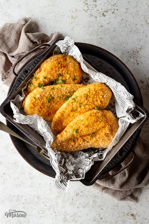 4 x Juicy air fried chicken breasts with a crispy golden crumb coating in a baking tray lined with baking paper. Set over a grey pewter tray on a light brown linen napkin. There is also a fork on the side.