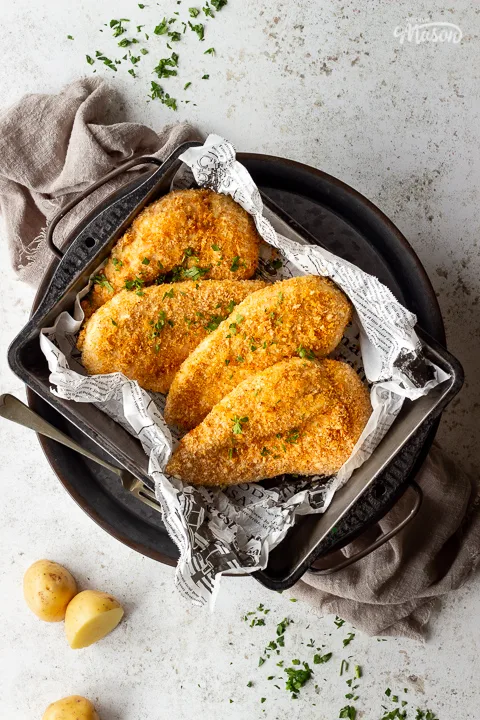4 x Juicy air fried chicken breasts with a crispy golden crumb coating in a baking tray lined with baking paper. Set over a grey pewter tray on a light brown linen napkin. There is also a fork on the side and some scattered potatoes and chopped parsley in the background.