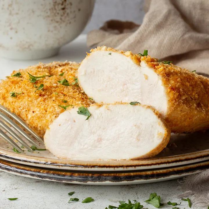 A close up of a sliced juicy air fryer chicken breast on a plate with a fork. Set against a mottled white backdrop.