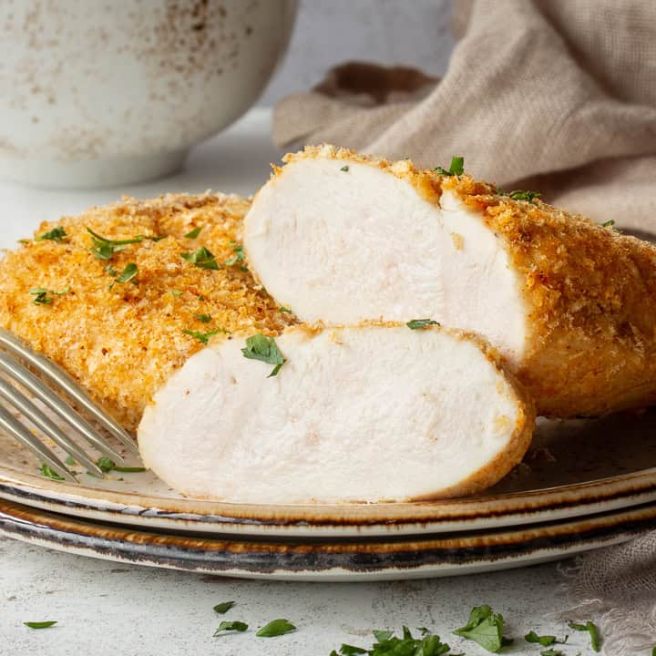 A close up of a sliced juicy air fryer chicken breast on a plate with a fork. Set against a mottled white backdrop.