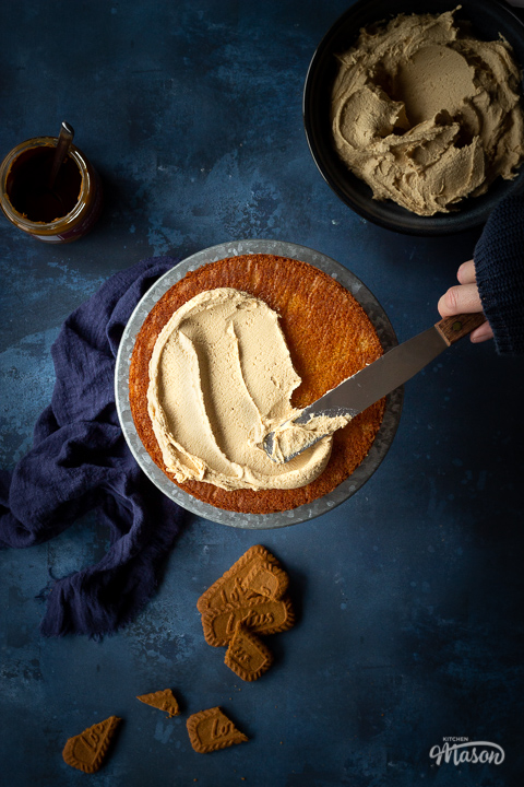 A vanilla sponge being coated with Biscoff frosting, against deep blue backdrop.