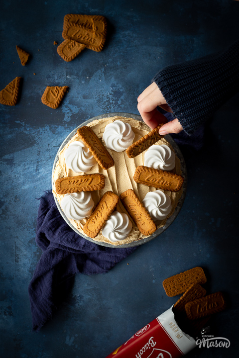 Top down view of a Biscoff cake on a cake stand being decorated with Biscoff biscuits and mini meringues. Set against a deep blue backdrop with broken Biscoff biscuits scattered in the background.