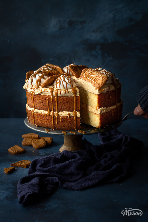 Someone taking a slice of Biscoff cake using a cake slice. The remaining cake is on a cake stand with a blue linen napkin strewn around its base.