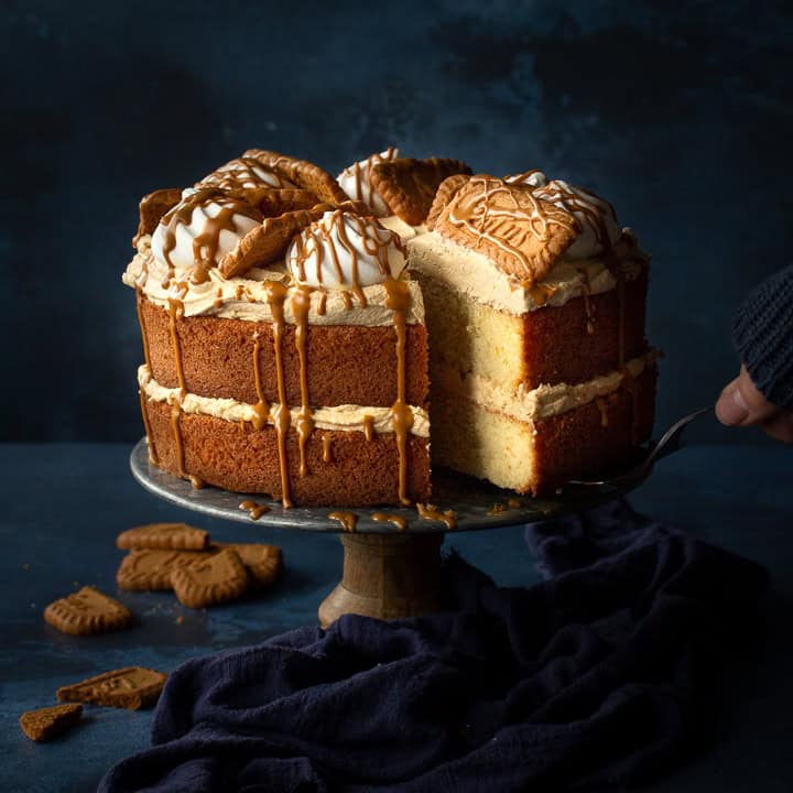 Someone taking a slice of Biscoff cake using a cake slice. The remaining cake is on a cake stand with a blue linen napkin strewn around its base.