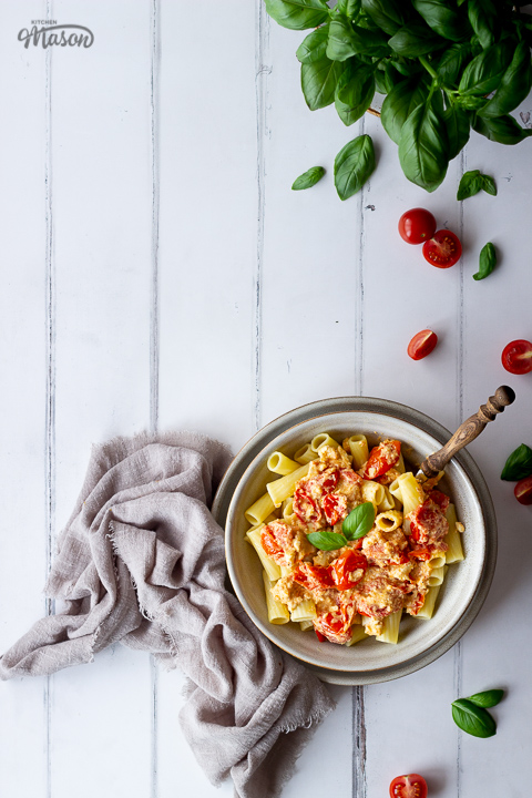 A bowl of tomato feta pasta surrounded by basil and chopped tomatoes