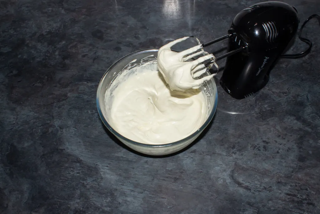 Whipped sweetened vanilla cream in a glass bowl with an electric hand whish