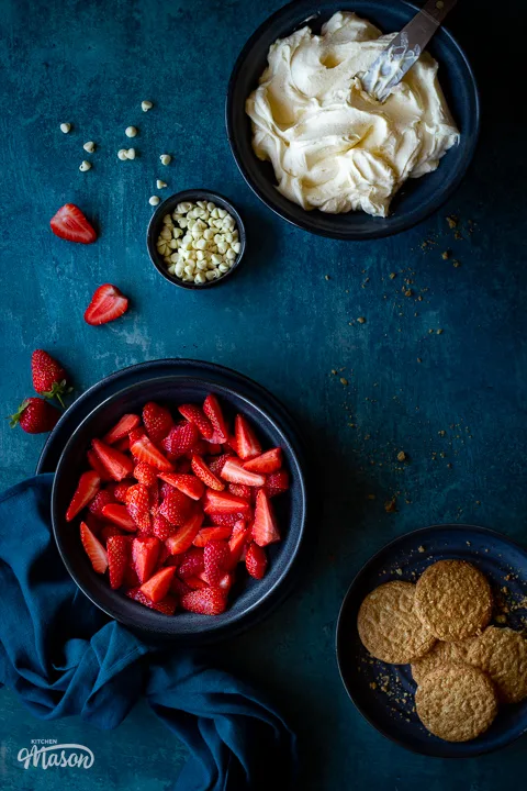 Chopped strawberries in a bowl, sweetened whipped cream in a bowl and some biscuits on a plate