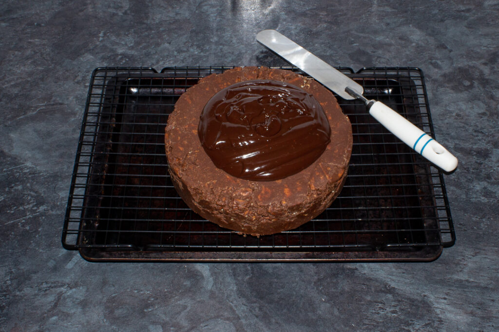 Chocolate ganache being smoothed over a biscuit cake on a wire rack