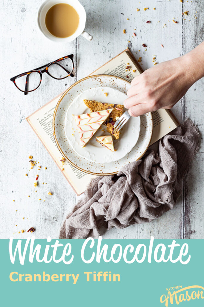 Someone forking a slice of white chocolate tiffin on a plate over a book. A text overlay says White Chocolate Cranberry Tiffin.