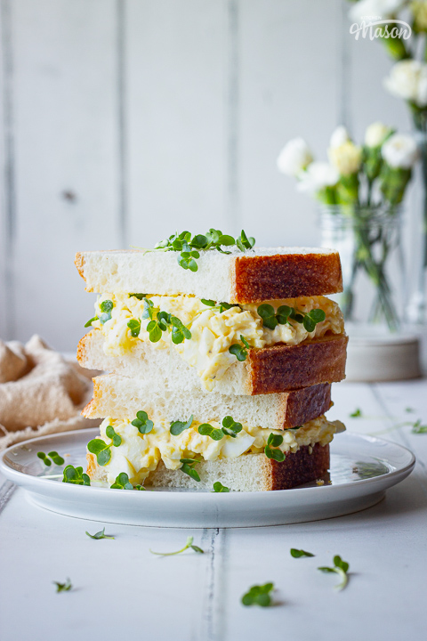 An egg salad sandwich on a plate with a vase of flowers