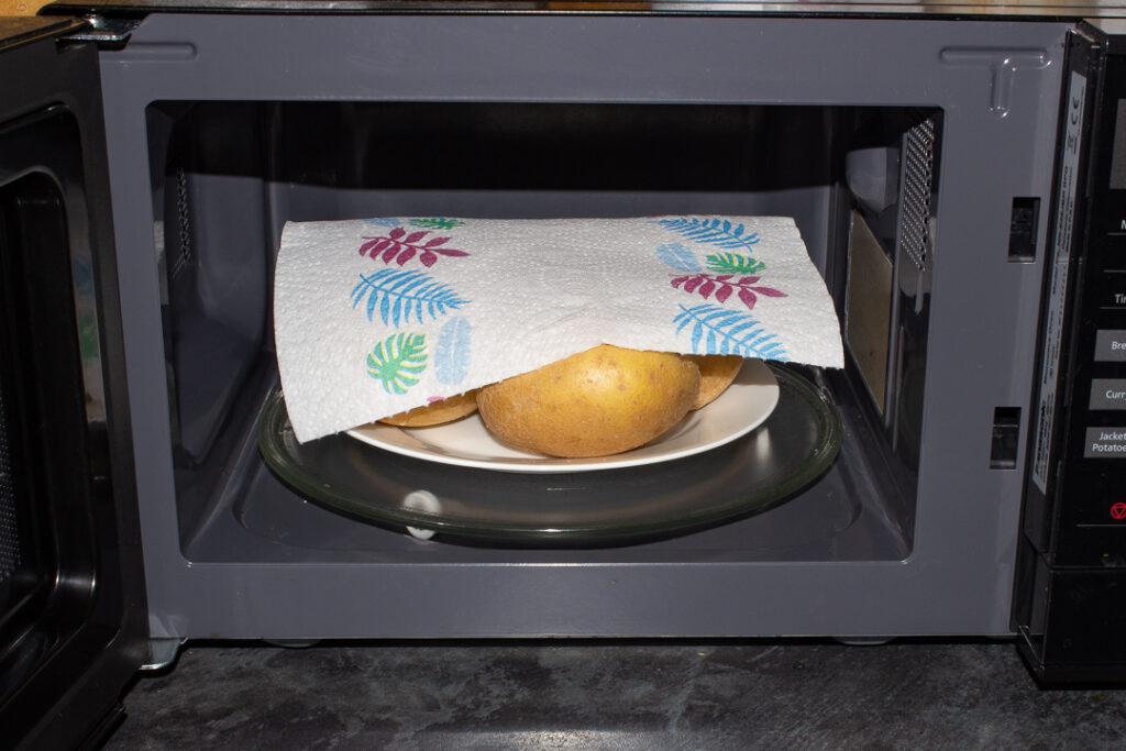 4 potatoes on a plate, covered with kitchen roll, in a microwave.