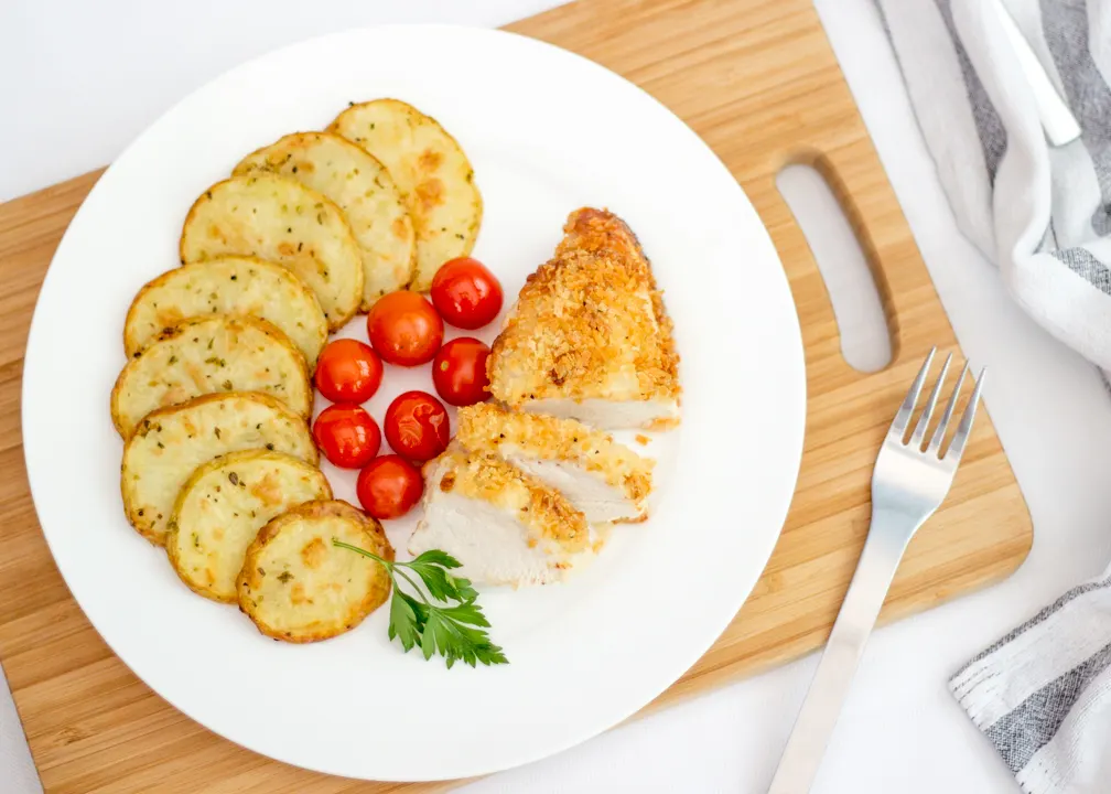 Baked parmesan chicken, potato thins and tomatoes on a plate with a fork on the side