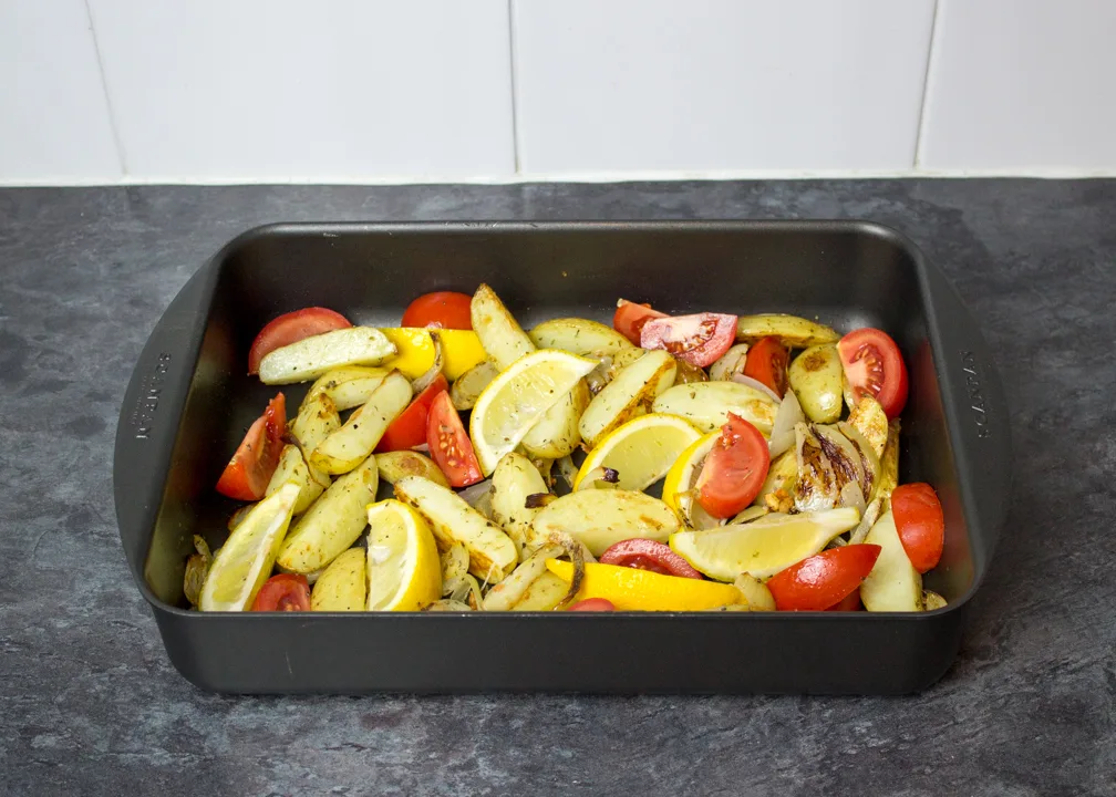 Partially cooked potato wedges with lemon and tomato wedges in a roasting dish