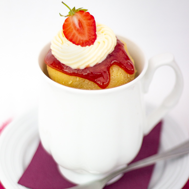Vanilla mug cake in a white mug topped with strawberries and sweetened cream.