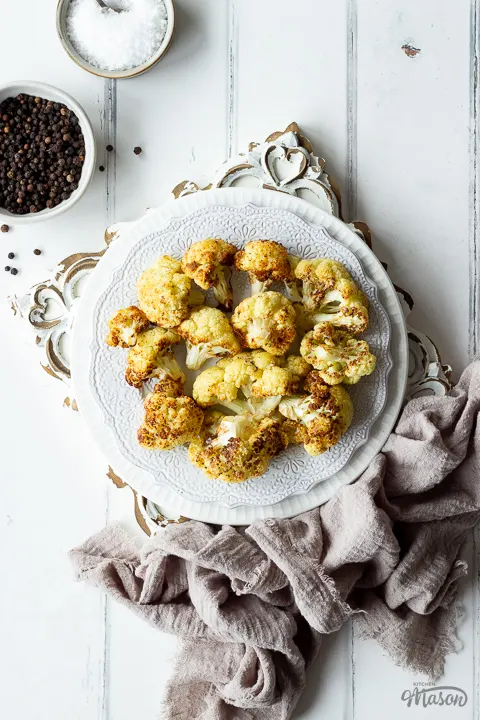 Close up of roast cauliflower on a white plate with pots of peppercorns and salt on the side