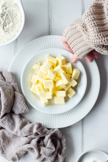 Someone putting a plate of cubed butter down next to flour, eggs and sugar.