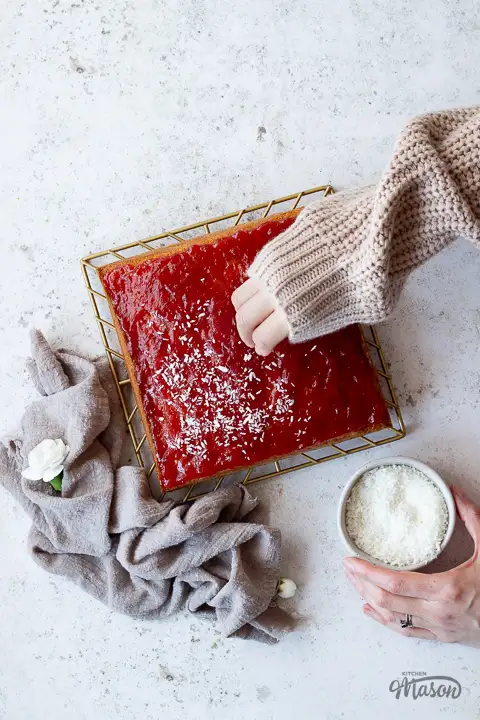 Coconut being sprinkled over a jam topped cake.