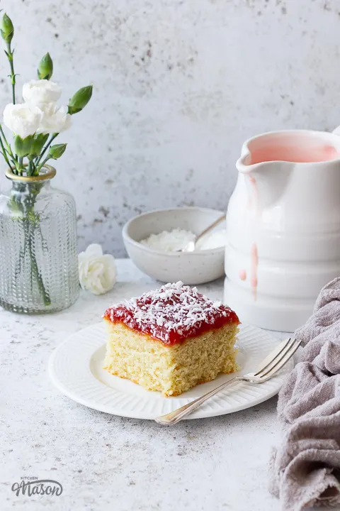 A slice of coconut jam cake on a plate with a jug of pink custard.