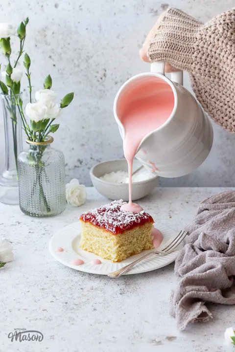 Pink custard being poured over a slice of coconut jam cake.