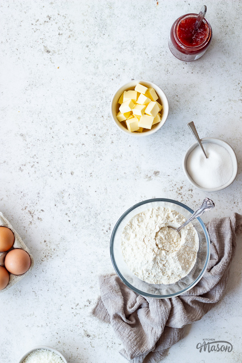 Flour, butter, sugar, jam and eggs in bowls.