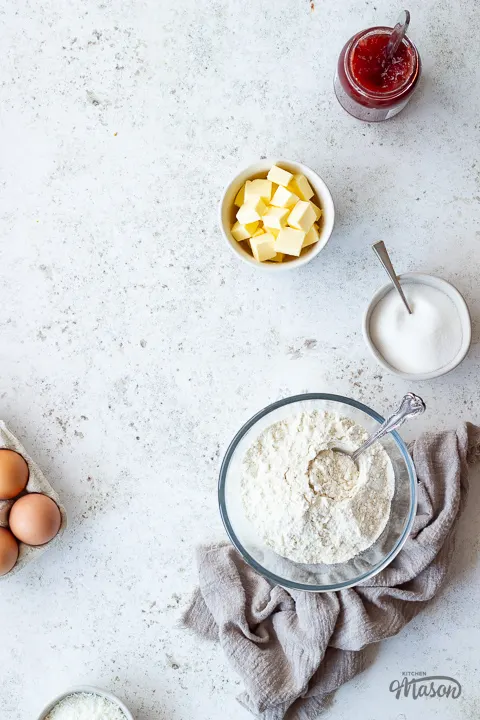 Flour, butter, sugar, jam and eggs in bowls.