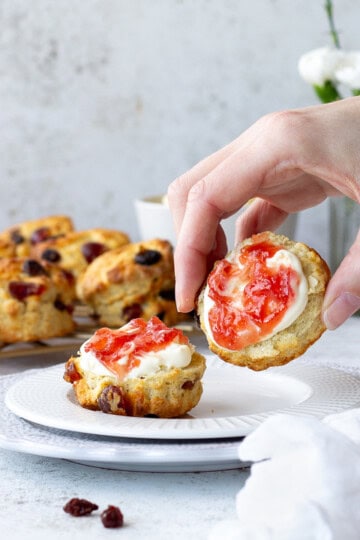 Someone picking up a fruit scone topped with jam and clotted cream.