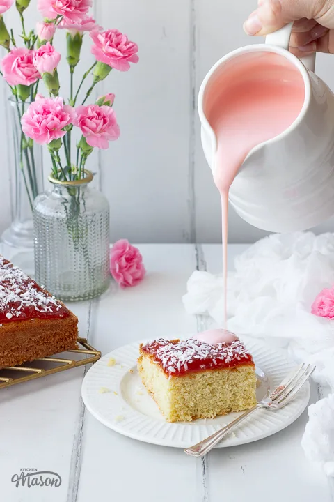 Pink custard being poured over coconut jam cake