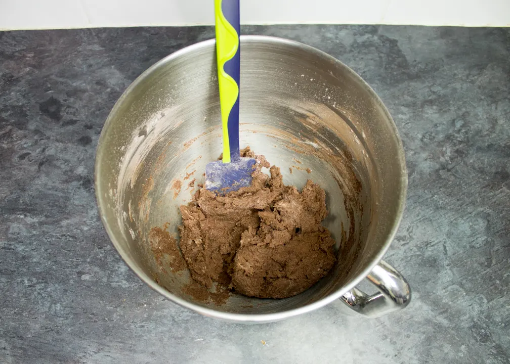 Chocolate biscuit dough in the bowl of an electric stand mixer with a spatula.