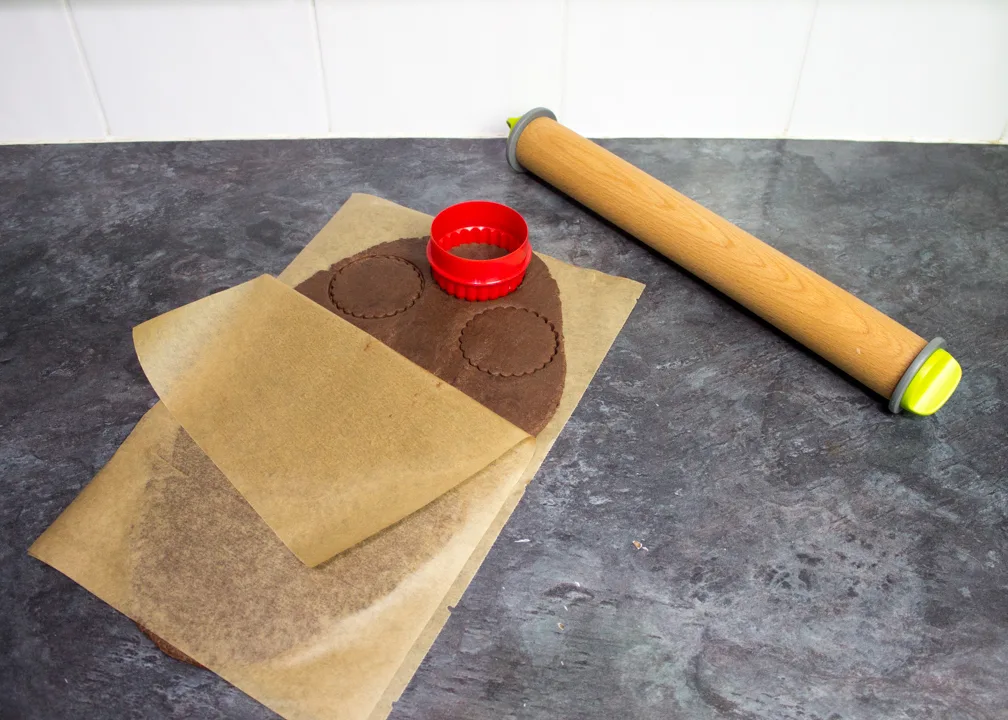 Chocolate biscuit dough rolled out between two sheets of baking paper, being cut into 7cm biscuits.