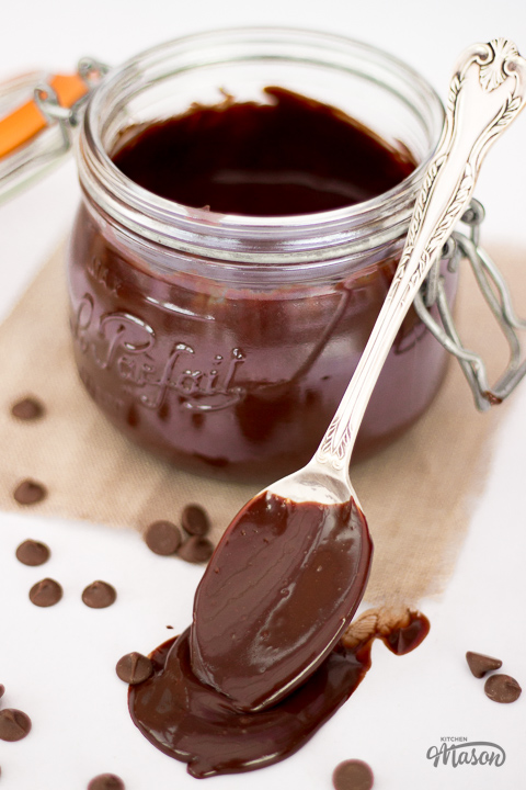 A jar of chocolate sauce with a spoon resting against it. The spoon is covered in chocolate sauce.