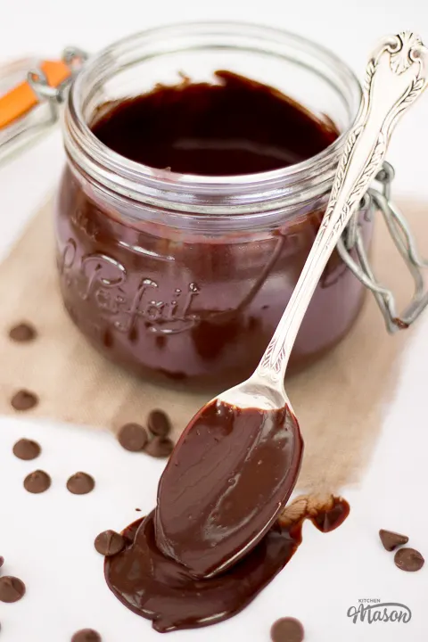 A jar of chocolate sauce with a spoon resting against it. The spoon is covered in chocolate sauce.