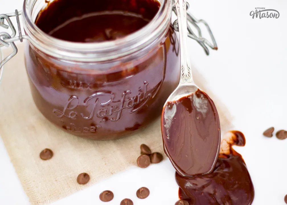 A jar of chocolate sauce with a spoon resting against it. The spoon is covered in chocolate sauce and there are chocolate chips scattered around it.