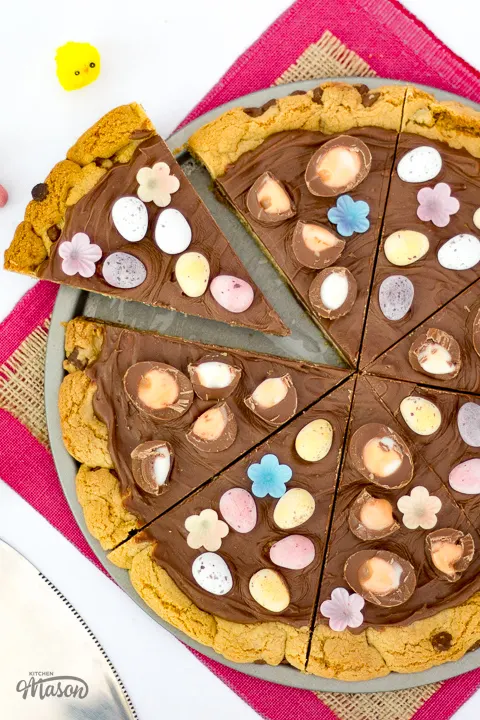 A sliced Easter chocolate cookie pizza on a baking tray with a cake slice next to it.