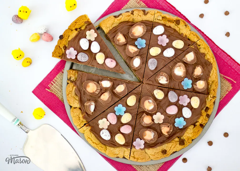 A sliced Easter chocolate cookie pizza on a baking tray with little yellow ornamental chicks and a cake slice next to it.
