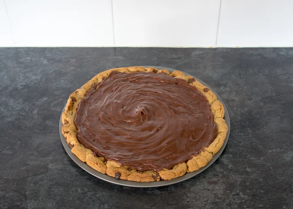 A chocolate chip cookie pizza on a baking tray topped with a chocolate ganache.