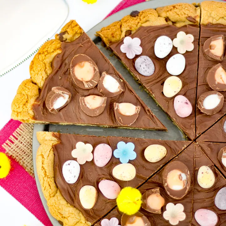 A sliced Easter chocolate cookie pizza on a baking tray with little yellow ornamental chicks.