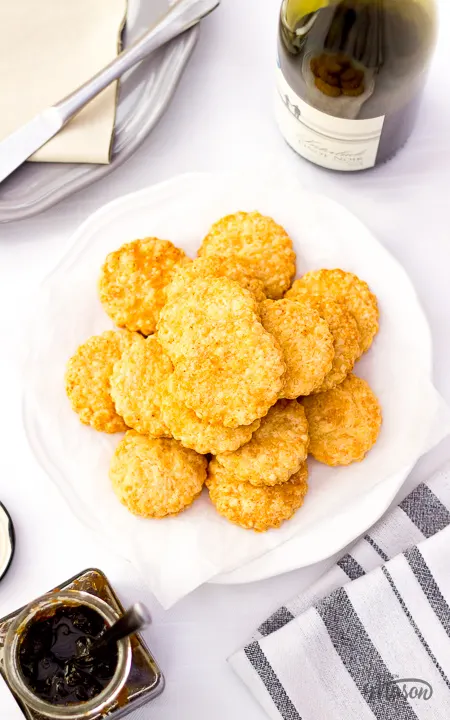 A white plate filled with lots of cheese biscuits. There's a bottle of wine and a jar of chutney to the side.