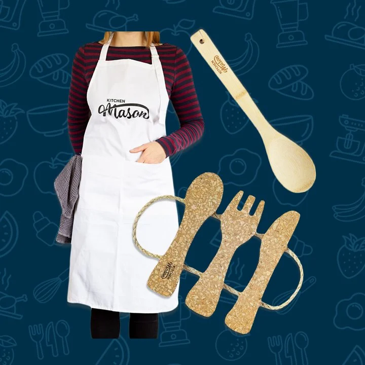 A Kitchen Mason branded white apron, cork pot stand and bamboo wooden spoon against a blue background.
