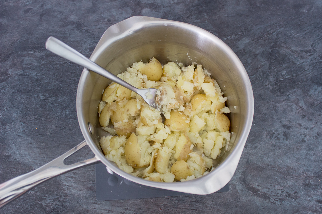 Potatoes being crushed with a fork in a saucepan.