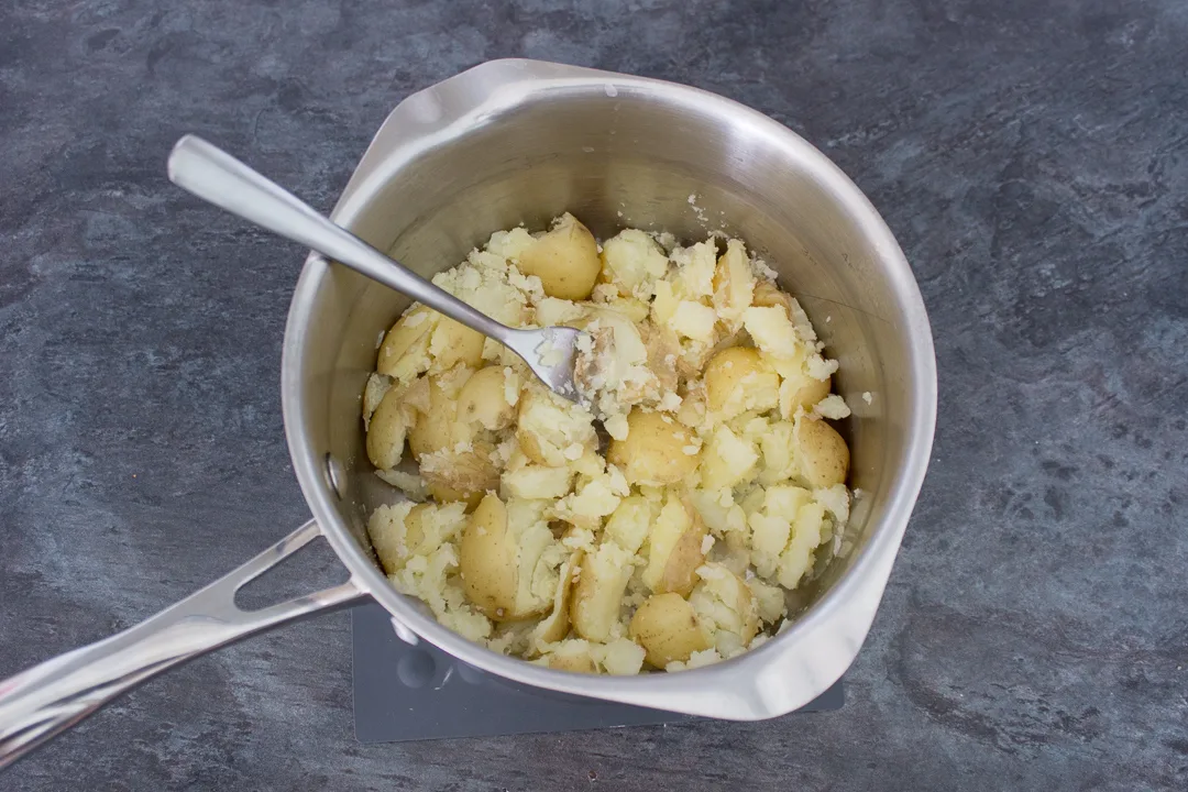 Potatoes being crushed with a fork in a saucepan.