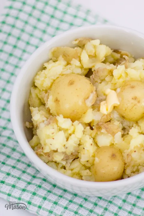 Crushed new potatoes in a white bowl over a green gingham tea towel.